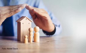 von zwei Händen in Dachform geschütztes Holzhaus mit Holzfiguren-Familie