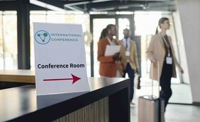 Ein Schild weist auf den Konferenzraum in einem Hotel. Im Hintergrund sind Teilnehmer zu sehen.
