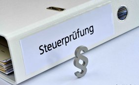 Weißer Aktenordner mit der Aufschrift "Steuerprüfung" liegt auf einem Tisch. Davor steht ein graues Paragrafenzeichen.