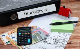 Ordner mit Aufschrift Grundsteuer, Taschenrechner, Kugelschreiber, Notizblock, Geldscheine, Grundrissplan
